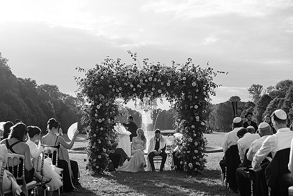Outdoor wedding ceremony as guests watch the Jewish wedding ceremony under a floral chuppah on a lawn with fountain and trees in back