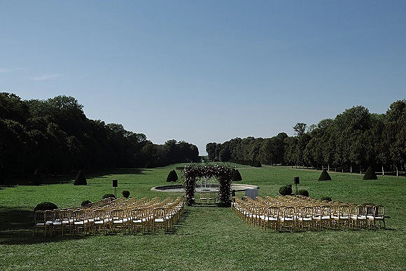 Outdoor ceremony setup with a floral arch and gold chairs in rows along a wide aisle, set on a manicured lawn by a fountain under blue sky