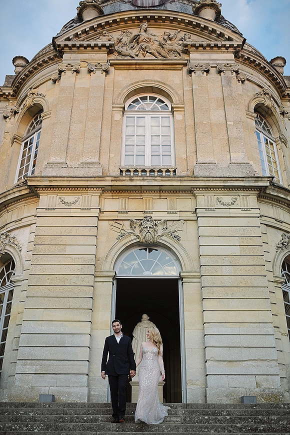 Couple portrait of bride and groom holding hands on a grand staircase, her beaded gown sparkling beside a stone facade and arched doorway