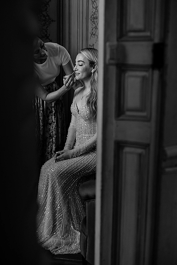 Bridal makeup being applied as the bride wears a beaded wedding dress and drop earrings in an interior room by a paneled door