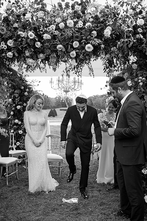 Ceremony moment as bride and groom stand at altar under a floral arch with roses and greenery on an outdoor lawn beneath a chandelier