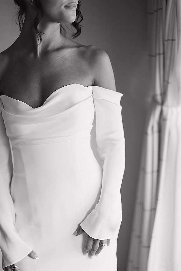 Wedding dress with an off the shoulder neckline and long sleeves, pearl earrings and ring detail, lit by soft window light and curtains indoors