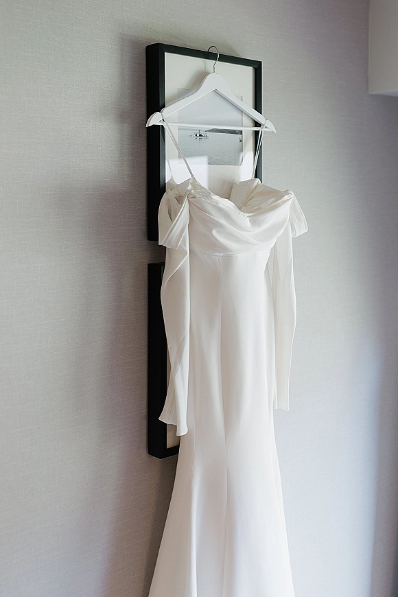 Wedding dress on a white hanger, minimalist wedding dress with spaghetti straps, draped neckline and off-shoulder sleeves in an indoor room
