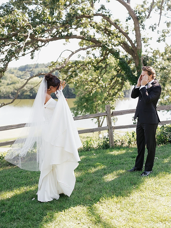 First look moment outdoors as bride in long veil taps groom’s shoulder by a lakeside wooden fence under tree branches