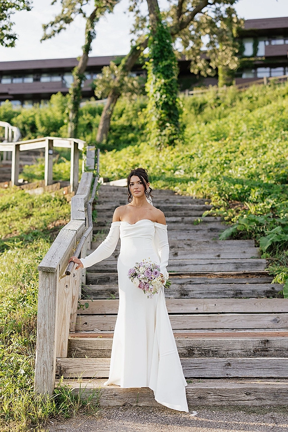 Bridal portrait of a bride in an off the shoulder wedding dress holding a purple flower and greenery bouquet on wooden steps by ivy-covered building