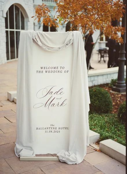 Wedding welcome sign with calligraphy lettering on an easel, draped with linen fabric in a stone courtyard with fall foliage