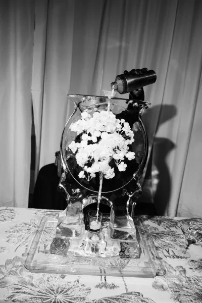 Ice sculpture wedding ice sculpture drink display with floral accent as a bottle pours into an ice bowl, surrounded by cocktail glasses on a draped table