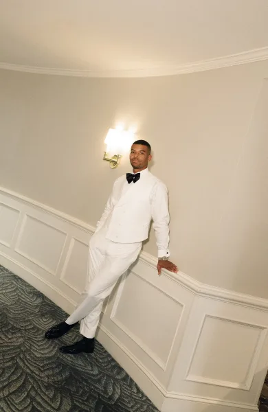 Groom portrait in a white tuxedo and black bow tie, leaning in an indoor hallway with paneled wall, cufflinks and watch visible