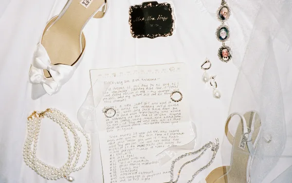 Wedding details flatlay with bridal detail flatlay of rings, pearl jewelry, veil, white bow heels, and handwritten vow letter on white fabric backdrop