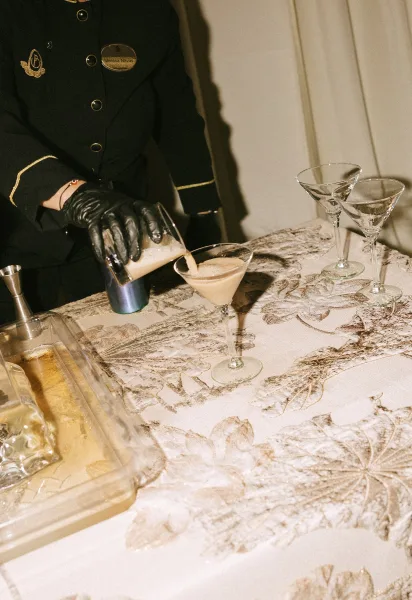 Wedding cocktail service with a gloved bartender pouring into three martini glasses on a tray at a bar table with wall paneling behind