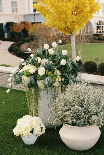 Ceremony floral arrangements with wedding aisle florals in tall urns of white hydrangeas and roses with hanging greenery along a lawn walkway