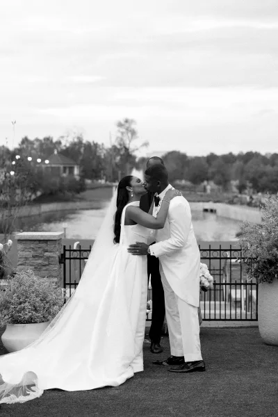 Wedding kiss portrait of bride and groom kissing, her long veil draped as he wears a white tuxedo, with lake and trees behind