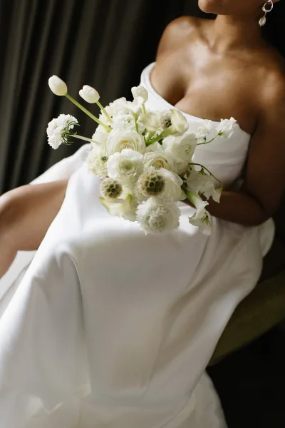 Bridal bouquet of ranunculus and tulips in an all-white round arrangement, held by a bride in a strapless dress before dark drapery