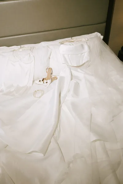 Wedding dress in a bridal details flat lay on a bed with veil, bow heels, pearl bracelet, and lace lingerie against pillows