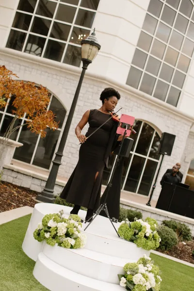 Wedding ceremony musician, wedding violinist in a black dress playing violin on a small outdoor riser beside white florals and a stone facade