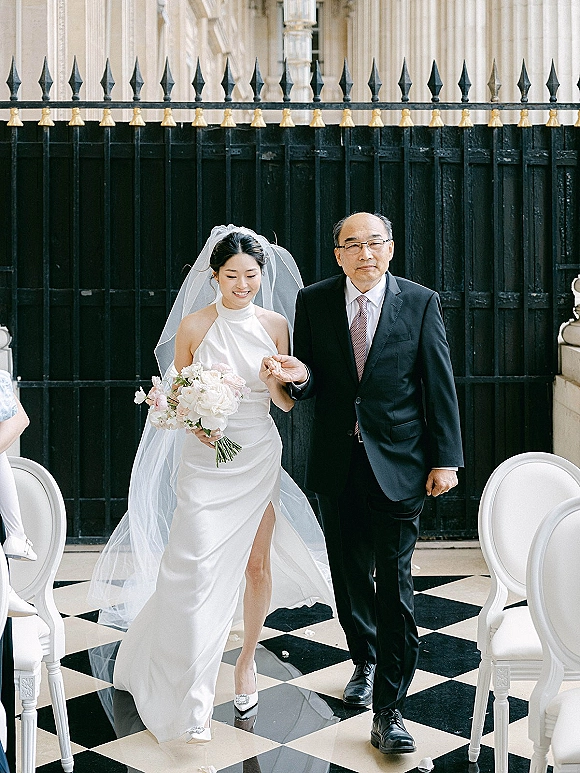 Bride walking aisle with bouquet and long veil, escorted by father in suit, past black iron gate on checkered tile floor