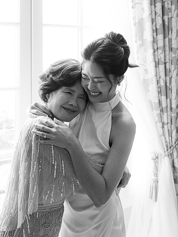 Bride hugging mother in window light, showing a high neck wedding gown and mother’s beaded sequined shawl against soft curtains