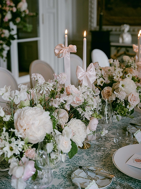 Reception tablescape with wedding table centerpiece of blush roses and peonies, taper candles with bows, and blue toile tablecloth place settings