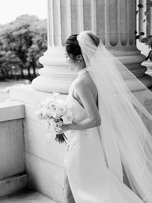 Bridal portrait of a bride in side profile holding a bouquet, long veil flowing beside stone columns with trees behind