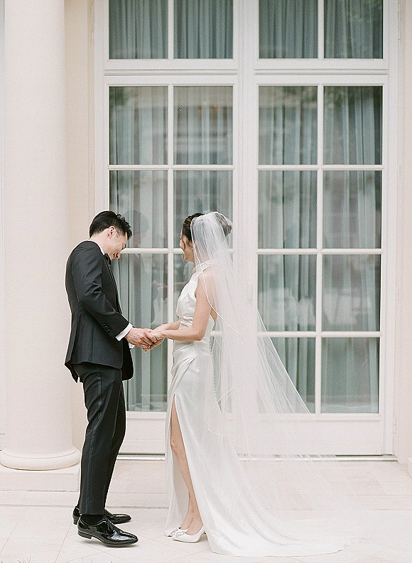 Couple portrait of bride and groom holding hands, bride in cathedral veil and satin dress beside large window panes and white columns