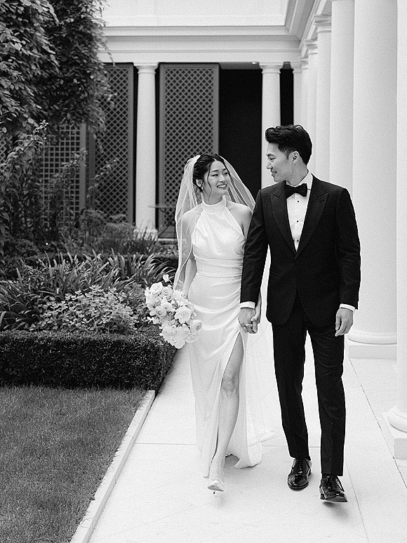 Couple portrait of bride and groom walking hand in hand, she holds a white rose bouquet and veil in a columned garden walkway