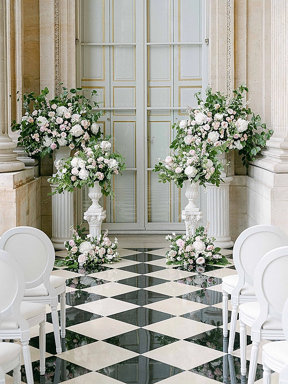 Ceremony altar flowers in tall wedding floral urns with roses, peonies and greenery on pedestals beside paneled doors on a checkered floor.