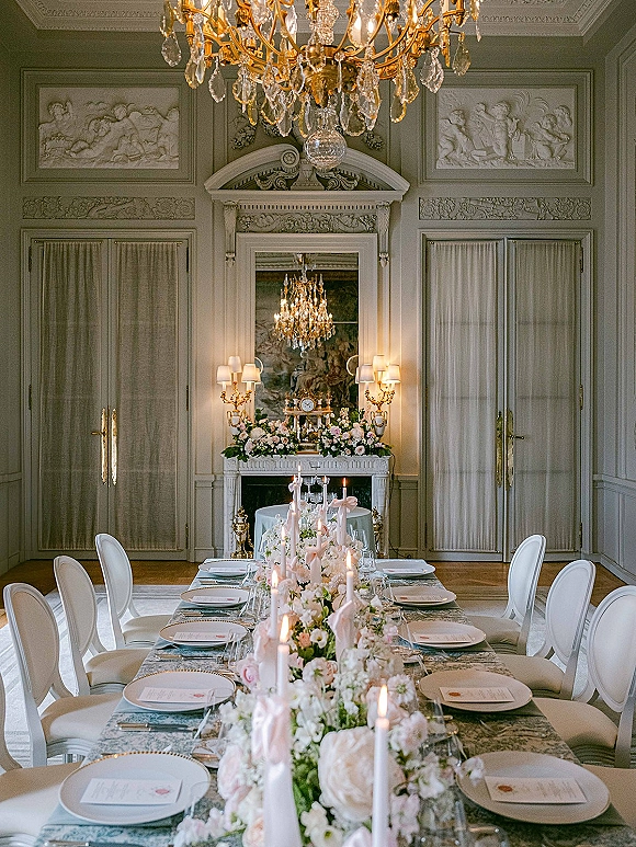 Reception tablescape with a long banquet table wedding setup, floral garland runner and taper candles beneath a crystal chandelier in an ornate dining room