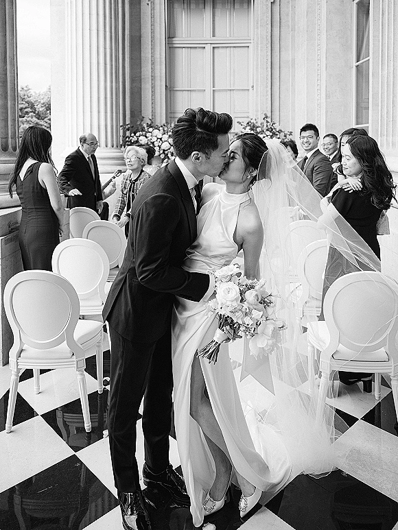 Wedding kiss portrait of bride and groom kissing on a checkered aisle, bride holding bouquet with long veil, guests and columns behind