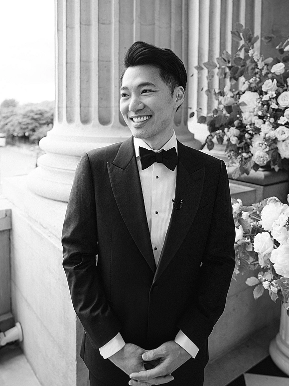 Groom portrait in a black tuxedo, hands clasped with a boutonniere, standing by stone columns on an outdoor terrace with florals