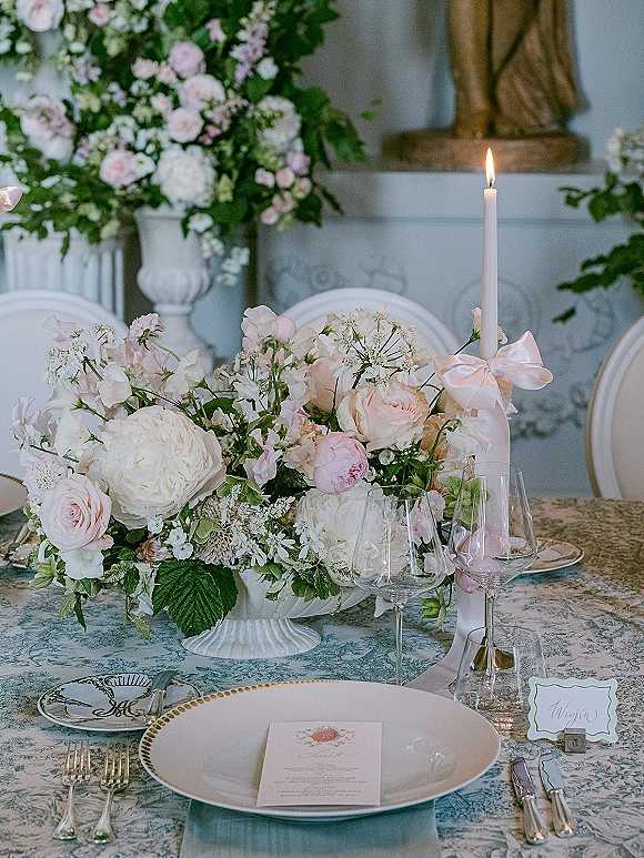 Reception tablescape with wedding table centerpiece in a white compote, blush roses, hydrangea, and taper candle with pink ribbon bow on blue toile linen