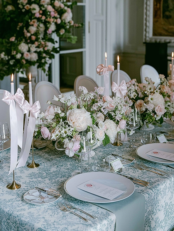 Reception tablescape with a wedding table centerpiece of blush and white florals, taper candles with pink bows, and formal place settings in a bright room