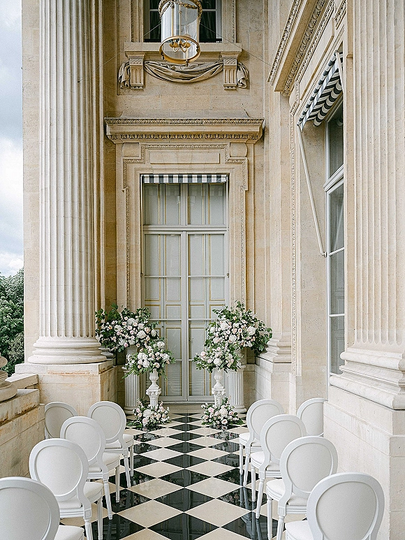 Ceremony setup with outdoor ceremony aisle, white chairs and floral pedestal urns on a checkered terrace before stone columns and doors