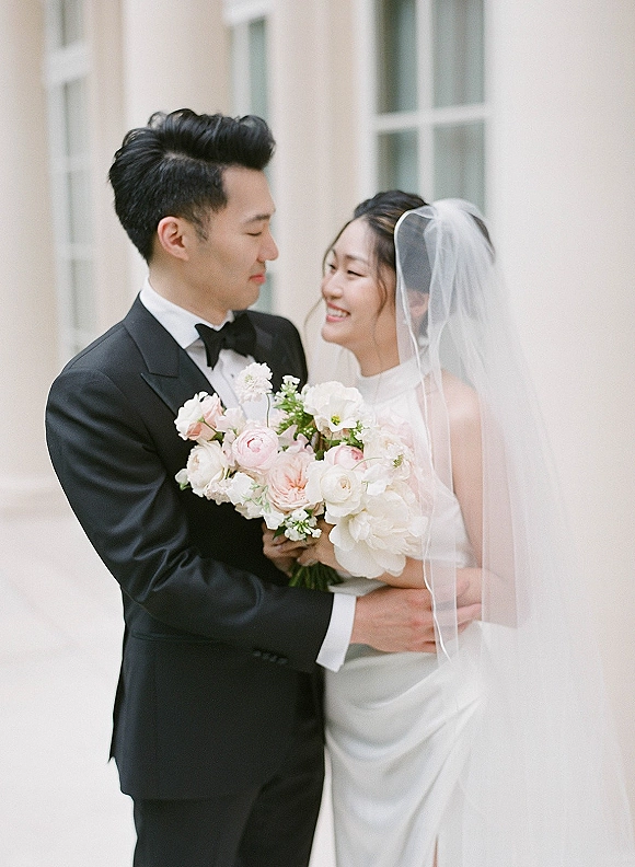 Couple portrait of bride and groom portrait hugging, her veil and bouquet beside his black tuxedo, against a pale wall and window backdrop