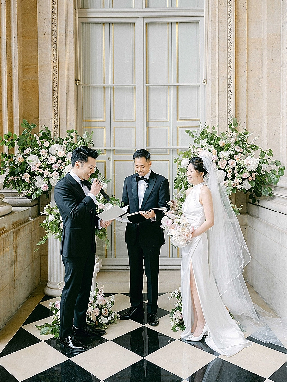 Wedding ceremony moment as groom reading vows into a microphone beside bride with bouquet and veil before ornate doors, roses behind