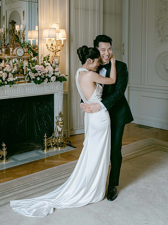 Couple portrait of bride and groom laughing as they hug by an ornate fireplace with floral mantel arrangement, clock, and candelabra lamps