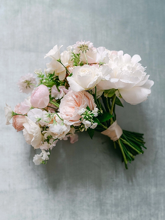 Bridal bouquet with white peonies and blush peonies, white roses, sweet peas, and greenery, tied with ribbon on gray fabric backdrop