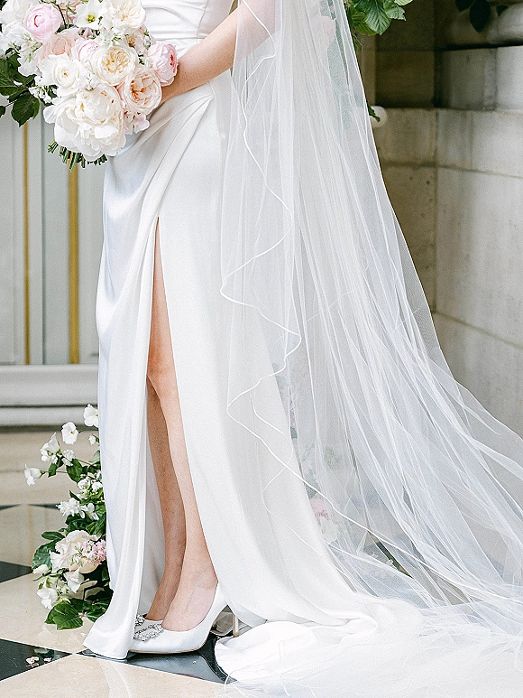 Bridal gown details on a satin wedding dress with thigh-high slit, veil and jeweled shoes beside a blush bouquet on marble steps indoors