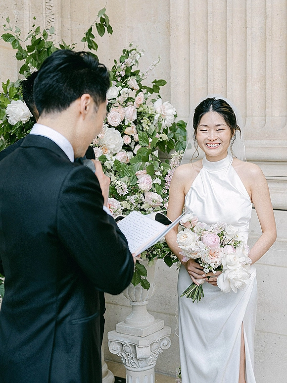 Wedding vows as groom reads from a vow booklet beside laughing bride with blush rose and peony bouquet, stone columns behind