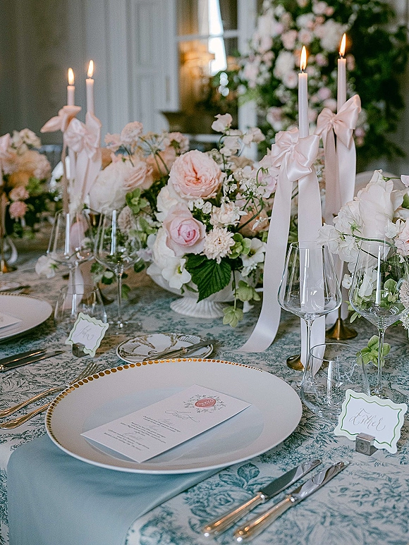 Reception tablescape with a wedding table setting featuring pastel garden rose centerpieces, bow taper candles, and patterned tablecloth in window light
