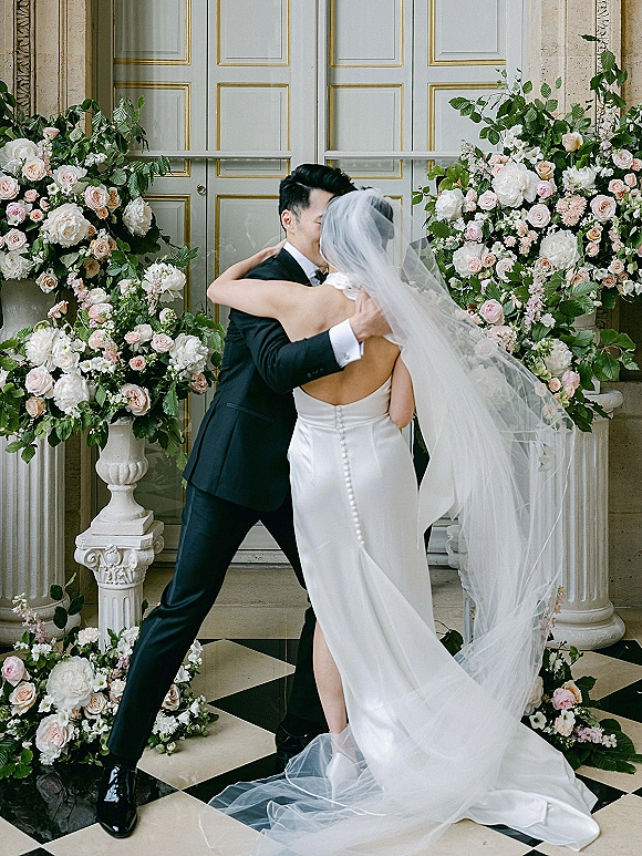 Wedding kiss portrait of bride and groom kissing beneath a veil, framed by columns and urn florals on a black-and-white floor