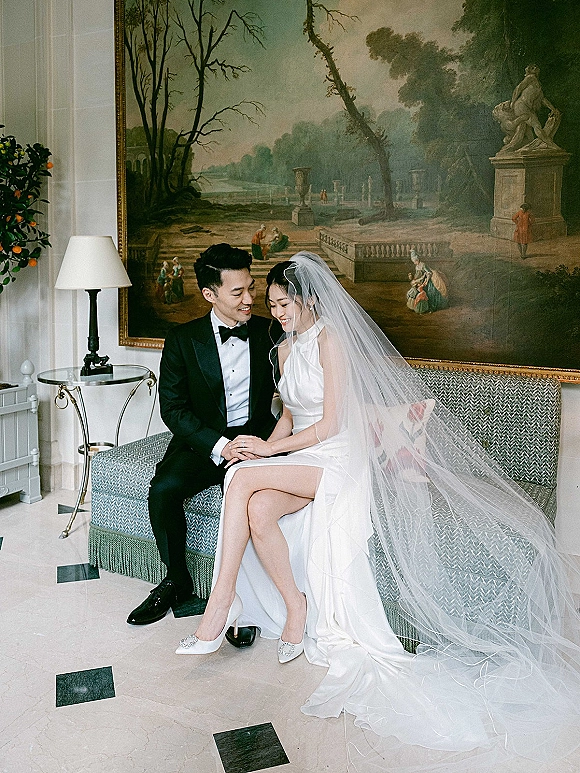 Couple portrait of bride and groom sitting on a sofa, holding hands as her cathedral veil drapes over marble floor in a lounge