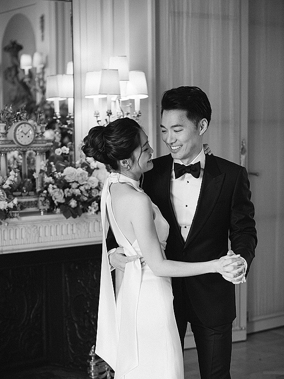 First dance as bride in a halter wedding dress and groom in tuxedo embrace by a fireplace mantel with flowers, lamps, and clock