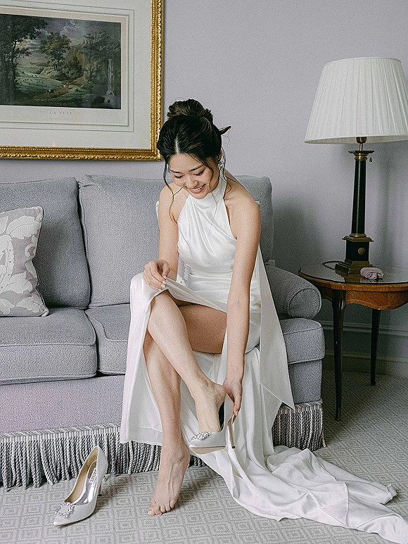 Bride getting ready, putting on wedding shoes with jeweled buckle heels while seated on a couch in a modern living room, hair in a sleek bun