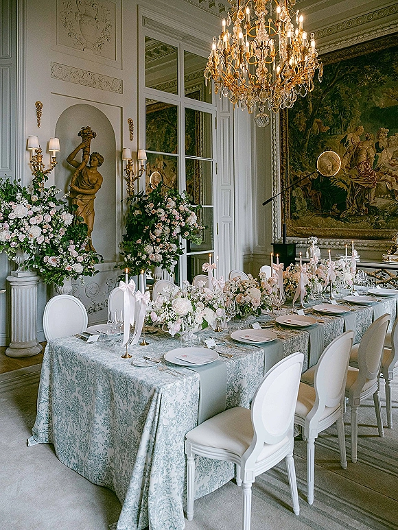 Reception tablescape with wedding banquet table featuring blue patterned linens, tall pink-white florals, and taper candles beneath a chandelier