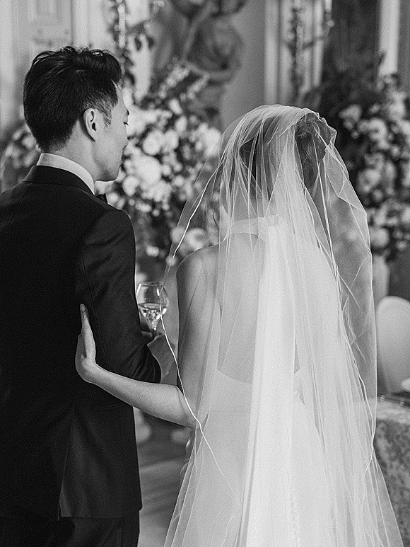 Ceremony moment as bride and groom from behind walk down the indoor aisle, her long veil trailing beside his black suit near altar flowers