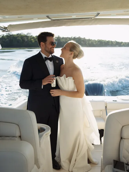 Couple portrait of a bride and groom on a boat, one wearing sunglasses, as the wake curls across open water with a forested shoreline behind.