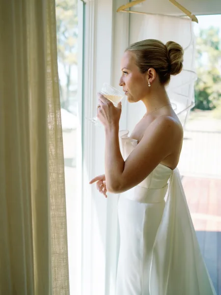 Bridal portrait of a bride with champagne, holding a coupe in a strapless satin gown by a bright window with curtains and trees beyond.