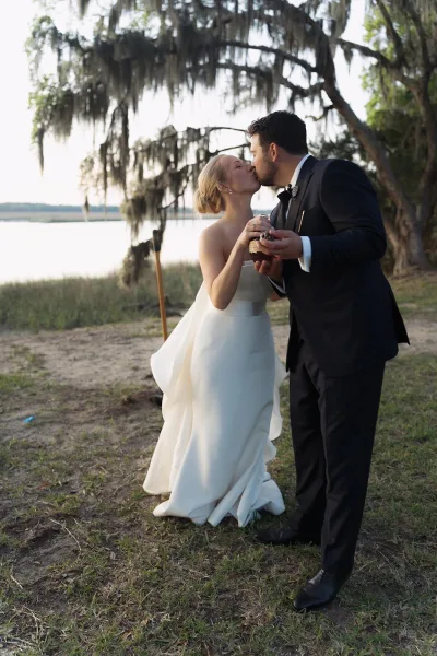Wedding kiss portrait in a lakeside wedding photo setting, the bride and the groom in a bow tie embrace beside a calm lake framed by trees.