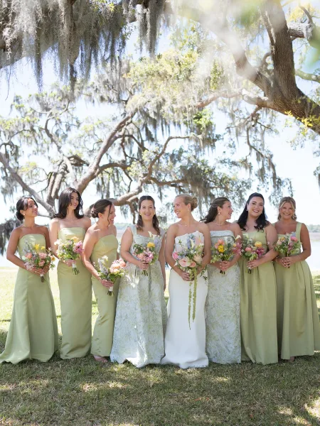 Bride and bridesmaids in sage green bridesmaid dresses holding bouquets beneath oak trees draped in Spanish moss beside a calm waterfront