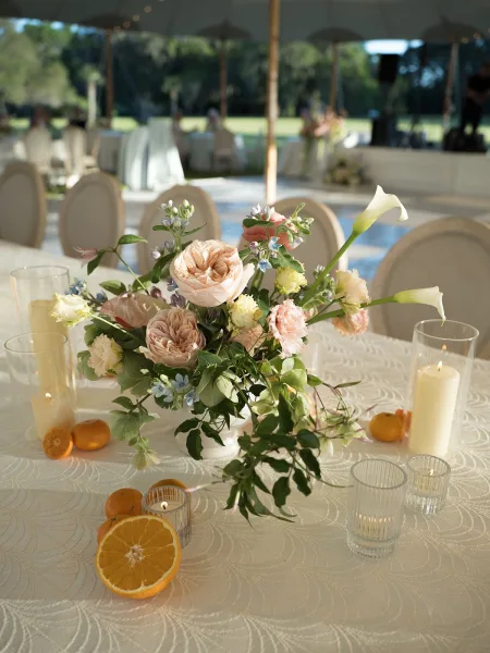 Wedding centerpiece featuring a citrus floral arrangement, accented by glass hurricanes on a tented lawn reception table.